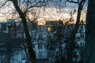 Günbatımının altın renkleri konut binalarının çatılarını aydınlatarak siluetli ağaçların dallarında sıcak bir parıltı oluşturur. Gökyüzü, yumuşak bulutlarla bezenmiş, gündüzden geceye geçiyor.