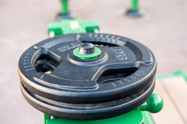 Ağır siyah bir plaka, açık hava fitness aktiviteleri için tasarlanmış yeşil bir egzersiz ekipmanına dayanır. Teçhizat sağlam ve bakımlı görünüyor, basit, işlevsel bir tasarım içeriyor.