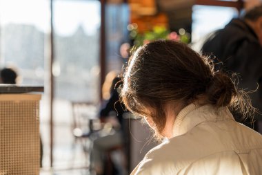 Sakin bir öğleden sonra, hareketli bir kafenin geniş pencerelerinden güneş ışığı akar. Saçları topuz şeklinde olan bir kadın sessizce oturur ve önündeki işe odaklanır. Sıcak, altın ışık