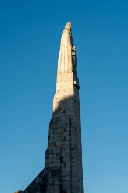 Karmaşık detaylarla oyulmuş yüksek bir taş heykel, açık mavi gökyüzüne ulaşır. Güneş ışığı, anıtların ihtişamını ve yüksekliğini vurgulayarak uzun bir gölge bırakır. Bu dönüm noktası
