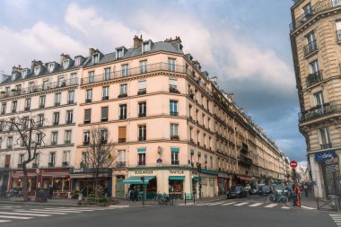 Paris 'in işlek bir sokak köşesinde büyüleyici mimari ve yerel işletmeler yer alıyor. Sahne, krem renkli duvarları ve balkonları olan klasik bir Paris binasını çekiyor. İnsanlar bisiklet sürerken görülüyorlar.
