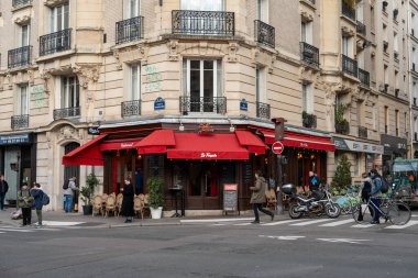 Canlı bir şehir ortamında, büyüleyici bir kafe, insanlar çeşitli etkinliklerle meşgul olarak yürürken kırmızı bir tenteye sahiptir. Açık hava oturma yerleriyle bu köşe kuruluşu hem yerel hem de ziyaretçileri çekiyor.