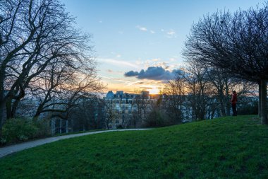 Huzurlu bir parkta, güneş ufkun altına batarken bireyler güzel günbatımının tadını çıkarırlar, manzarada sıcak bir parıltı bırakırlar. Uzun ağaçlar, kısmen kıştan kalma, resim çerçevesi