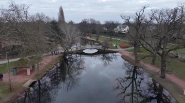 Sakin bir park sahnesinde bulutlu gökyüzünü yansıtan durgun bir göl sergileniyor. Çırılçıplak ağaçlarla çevrili büyüleyici bir köprü suyu kaplar. İnsanlar yollarda yürürler, sakin atmosferin tadını çıkarırlar. Şey...