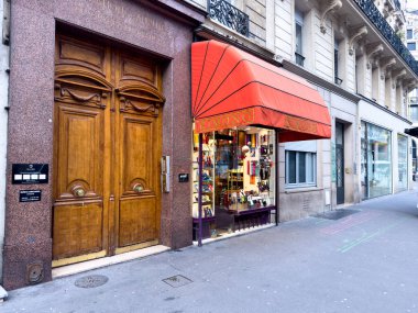 Paris, Fransa-19.02.2025 Parlak kırmızı tenteli bir dükkan çeşitli eşyalar sergiliyor, güneşli bir günde yoldan geçenleri çiziyor, zarif binalar caddeyi kaplıyor..