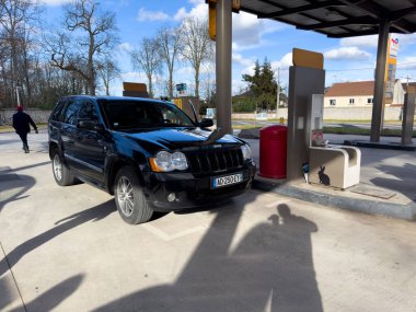 Dammarie-les-Lys, Fransa-23.02.2025 Siyah bir SUV benzin pompasının önünde dururken bir insan bulutlarla dolu bir gökyüzünün altında yürüyor ve sakin bir atmosfer yaratıyor..