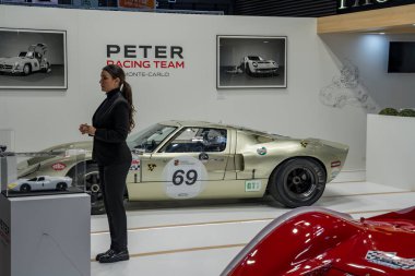 Paris,France-09.02.2025 Elegantly dressed presenter stands beside a classic racing car, engaging visitors with stories about its history at a Monte Carlo exhibition. clipart