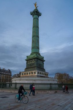Paris, Fransa-23.11.2024 Canlı bir şehir manzarası bisikletçilerin alacakaranlığın davetkar atmosferinde yayalar tarafından çevrelenmiş, yüksek yeşil bir sütunun yanından geçerken gözler önüne seriliyor.