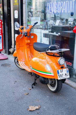 Paris, Fransa-20.20.2024 Bir göz alıcı turuncu scooter, yayaların ve yerel dükkanların öğleden sonranın coşkulu enerjisini somutlaştırdığı işlek bir caddenin vitrininde duruyor..