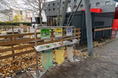 Paris, Fransa-03.12.2024 Geri dönüşüm için renkli çöp bidonları, serin bir sonbahar gününde şehrin kalbinde sürdürülebilir uygulamaları teşvik ederek, düşen yapraklar arasında sakin bir parkta oturuyorlar..