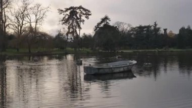 Bois de Vincennes 'de sakin bir gölü çevreleyen yemyeşil ağaçlar, akşam ışığında bulutların suyun üzerine yumuşak gölgeler düşürdüğü boş bir kayığın nazikçe yüzdüğü yer..