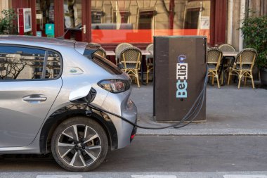 Şık bir elektrikli araç, güneşli bir öğleden sonra, modern şehir yaşamını gözler önüne seren, cafe koltukları ve yeşillikle kaplı bir kafenin önünde şarj edilir..