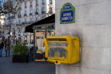 Taştan bir duvara eklenmiş renkli posta kutusu, kalabalık bir cadde Gambetta sabahında yürüyen insanlar, şehir cazibesini ve günlük gezintileri sergiliyorlar..