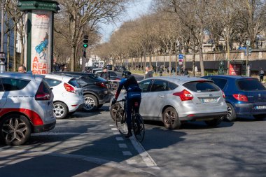 Bisikletçiler, açık mavi bir gökyüzü ve işlek caddeleri aydınlatan güneş ışığı altında, arabalarla ve yayalarla dolu canlı bir şehir kavşağında yol alırlar..