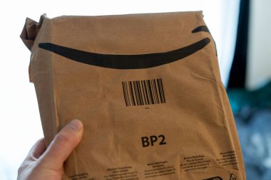 Bir el, gülümseyen bir logoyla süslenmiş karton bir paketi kavrıyor. Güneş ışığı sıcak bir odanın penceresinden içeri girerken heyecanını açığa vuruyor..