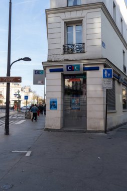 İnsanlar canlı bir Paris caddesinde, modern bir bankanın önünden geçiyor. Güneş, şehir aktiviteleriyle dolu canlı bir sahnede ışıldıyor..