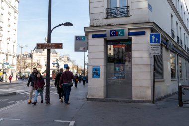 Yayalar hareketli bir kaldırımda yürürler, açık bir gökyüzünün ve sıcak güneş ışığının keyfini çıkarırken, canlı bir şehir ortamında dükkanların ve bir bankanın önünden geçerler..