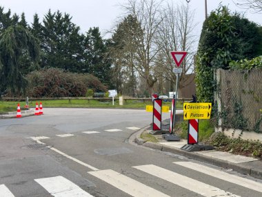 Villennes-sur-Seine, Fransa-26.02.2025 Parlak sarı işaretler düz yayaları güvenli bir şekilde gösterirken, yol çalışmaları yumuşak gri gökyüzü ve çevreleyen yeşillikler arasındaki kavşakta değişikliklere yol açıyor..