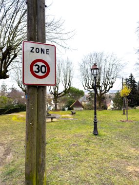 Villennes-sur-Seine, Fransa-26.02.2025 Bir antika park 30 km saat hız sınırını gösteren bir tabela sergiliyor, çıplak ağaçlar ve sakin bir arkaplana karşı klasik bir lamba direği.