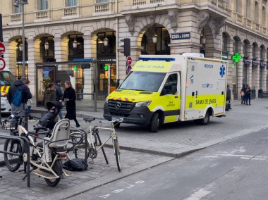 Kalabalık bir Paris caddesinde bir acil durum aracı park edilmiş durumda. Müdahale ekipleri, sakin bir sabah havasında vatandaşlara yardım ederken, şehir yaşamını ön plana çıkarıyor..