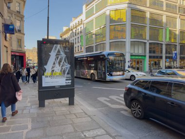 Paris, Fransa-05-03-2025 Bir şehir otobüsü güneşli bir günde trafikte yol alırken kaldırımda yürüyen yayalarla hareketli bir sokak sahnesi.