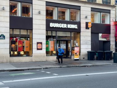 Paris, Fransa-08-03-2025 işlek bir şehir caddesinde renkli afişleri olan hareketli bir fast food müessesesi var. Yalnız bir müşteri günlük kıyafetlerini giyer ve düşüncelerine dalmış bir şekilde yürür..