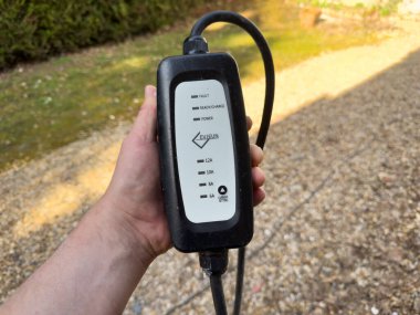 Villennes-sur-Seine, Fransa-08-03-2025 Huzurlu bir banliyö bahçesinde güneşli bir yolda dikilirken elektrikli araç şarj ünitesine sahip olan bir insan,.