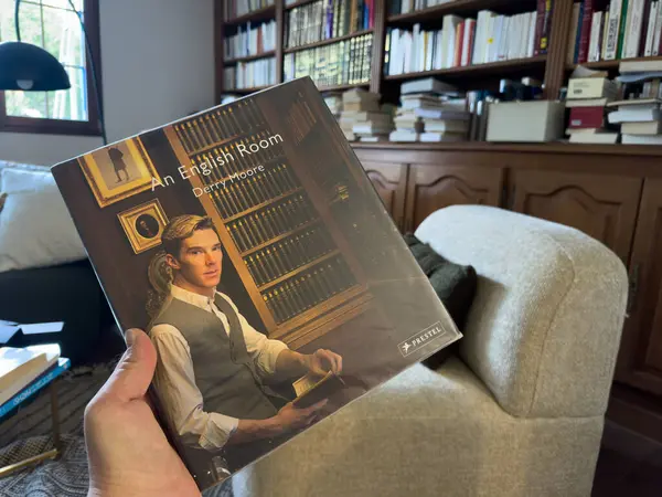 Villennes-sur-Seine, Fransa-12-03-2025 İyi giyimli bir birey, klasik edebiyat ve rahat mobilya rafları ile çevrili, rahat bir şekilde otururken bir İngiliz Odası başlıklı bir kitap tutuyor..