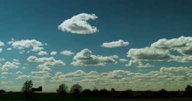 Birçok tüylü kümülüs bulutunun zamanı karanlık ufukta ağaçlarla ve binalarla birlikte mavi gökyüzünde dönüşüyor ve hareket ediyor.