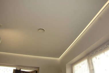 Pürüzsüz bir ışık yüzeyi olan esnek bir tavan. Tavan boyunca uzanan uzun ince şeritli şerit sıcak ve dağınık bir parıltı sağlıyor. Tavanın ortasında yuvarlak bir havalandırma görünür. Sağ alt köşede bir pencere kısmen görülebilir