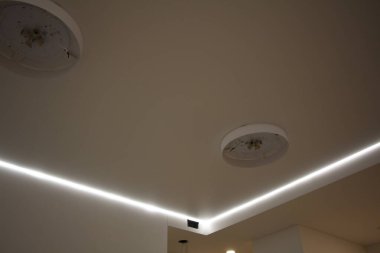 Pürüzsüz bir ışık yüzeyi olan esnek bir tavan. Tavan boyunca uzanan uzun ince şeritli şerit sıcak ve dağınık bir parıltı sağlıyor.. 