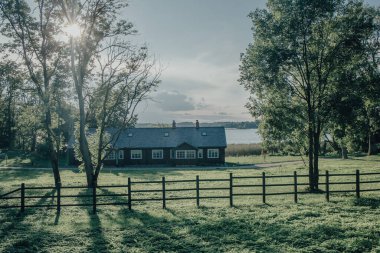 a house with a gray tile roof, located in lush greenery. The house is partially hidden by two tall trees in the foreground, and a rustic wooden fence runs along the bottom of the frame. In the background is a calm lake, reflecting soft sunlight. clipart