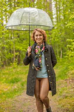 Bahar ormanında şemsiyeli genç bir kadın, muhtemelen yağmur yağmaya başladı, etrafta yeşil çalılar ve ağaçlar var..