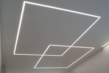 Eşsiz bir geometrik tasarımı olan modern bir tavan. Dahili LED şerit ışıklandırmayla yaratılmış. Işıklandırma birbirine kenetlenmiş iki kare şekil oluşturuyor, biri daha büyük ve biri daha küçük, pürüzsüz beyaz tavanda görsel olarak çarpıcı bir desen oluşturuyor..