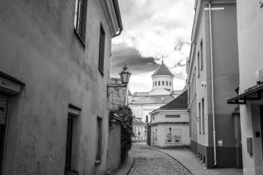 Siyah ve beyaz bir fotoğraf Vilnius 'taki sarmal kaldırım kaldırım kaldırımını (Skapo gatve) gösterir. Cadde pencereleri ve mimari detayları olan eski binalarla dolu..