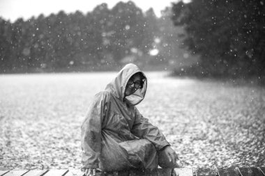 Kapüşonlu yağmurluk ve gözlüklü bir kadının tek renkli görüntüsü, bir göletin yanındaki ıslak tahtalarda oturuyor..