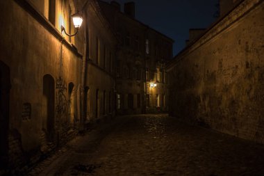 Vilnius Eski Kasabası 'ndaki karanlık ve dar bir patikanın yatay fotoğrafı. Sokağın her iki tarafında da taş duvarlar olan eski binalar yükseliyor. Ilık sarı ışığı olan birkaç eski fener sokağı aydınlatarak gizemli bir atmosfer yaratıyor..