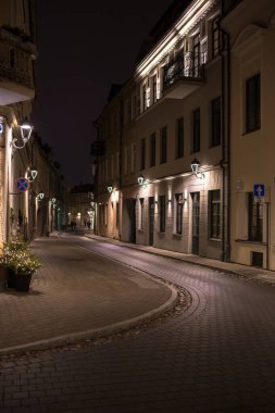 Vilnius Eski Kasabası 'ndaki dolambaçlı bir caddenin dikey fotoğrafı. Caddenin her iki tarafında da aydınlık pencereleri ve duvarlarında dekoratif fenerleri olan eski binalar var.. 