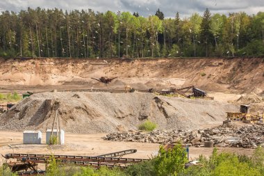 Teraslar ve makinelerle taş ocağının genel manzarası. Kazı makineleri ve diğer makineler taş ocağının farklı seviyelerinde çalışıyor. Zemin kırmızımsı-kahverengi bir renge sahiptir ve yeşil bitki örtüsü arka planda görünür..
