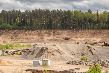 Teraslar ve makinelerle taş ocağının genel manzarası. Kazı makineleri ve diğer makineler taş ocağının farklı seviyelerinde çalışıyor. Zemin kırmızımsı-kahverengi bir renge sahiptir ve yeşil bitki örtüsü arka planda görünür..