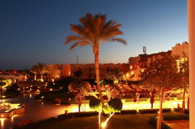Akşamın erken saatlerinde tatil köyü manzarası. Gökyüzü koyu mavi ve geleneksel otel binaları sıcak sarı bir ışıkla aydınlanıyor. Ön planda duran uzun bir palmiye ağacı.