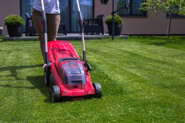 Kırmızı çim biçme makinesiyle çim biçen bir kadın. Çim biçme makinesi siyah elementlerle kırmızıdır. Çim biçme makinesinin arkasında yeşil bir çim ve çalıların yanı sıra arka planda ağaçlar görebilirsiniz. Güneşli bir gün, çimenler parlak yeşil.