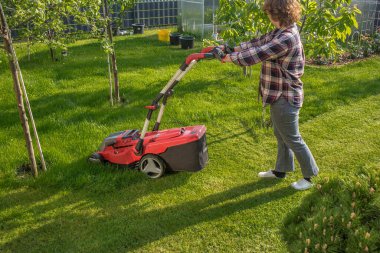 Kırmızı çim biçme makinesiyle çim biçen bir kadın. Çim biçme makinesi siyah elementlerle kırmızıdır. Çim biçme makinesinin arkasında yeşil bir çim ve çalıların yanı sıra arka planda ağaçlar görebilirsiniz. Güneşli bir gün, çimenler parlak yeşil.