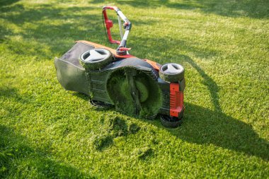 Kablosuz çim biçme makinesinin yakın çekimi. Çim biçme bıçağı görülebiliyor. Taze kesilmiş çimenler ve kırmızı vücudun bir parçası..