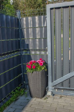 Modern bir çit ve kapının yanında parlak pembe ve kırmızı çiçekli uzun, koyu gri bir hasır ekici. Çit metal ağdan yapılmıştır..