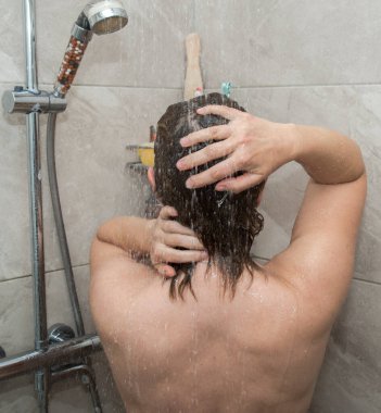 Duş kabininde duran birinin arkasından manzara. Saçını yıkayan kişi, saçında köpük var. Arka plan hafif bir fayans..