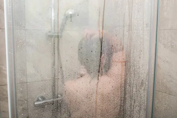 Duş kabininde duran birinin arkasından manzara. Saçını yıkayan kişi, saçında köpük var. Arka plan hafif bir fayans..