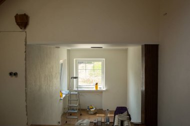 Tadilat yapılan bir oda. Ortasında kuşaklı bir pencere var. Pencerenin önünde bir merdiven var. Duvarlar boyamak için kısmen hazırlandı. Odada çeşitli yapı malzemeleri ve araçlar görülebilir.