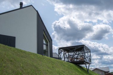 Minimalist tasarımı olan modern bir ev, yamaçta yer alıyor. Evin yanında, yamaçta, metal pergola ile açık bir çardak yapılır. Gökyüzü, kompozisyona dinamizm katan resimsel bulutlarla doludur..