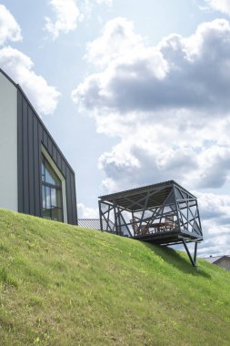 Minimalist tasarımı olan modern bir ev, yamaçta yer alıyor. Evin yanında, yamaçta, metal pergola ile açık bir çardak yapılır. Gökyüzü, kompozisyona dinamizm katan resimsel bulutlarla doludur..